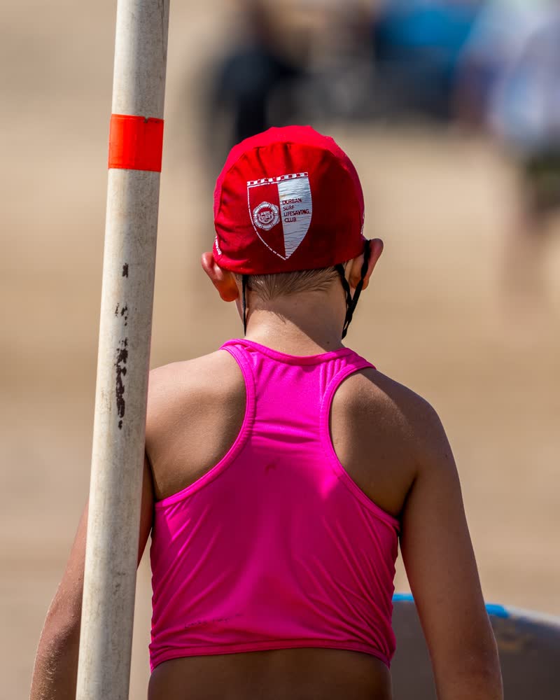 Young Durban Surf Lifesaving Club nipper in a red club cap and pink vest, seen from behind beside a beach marker pole